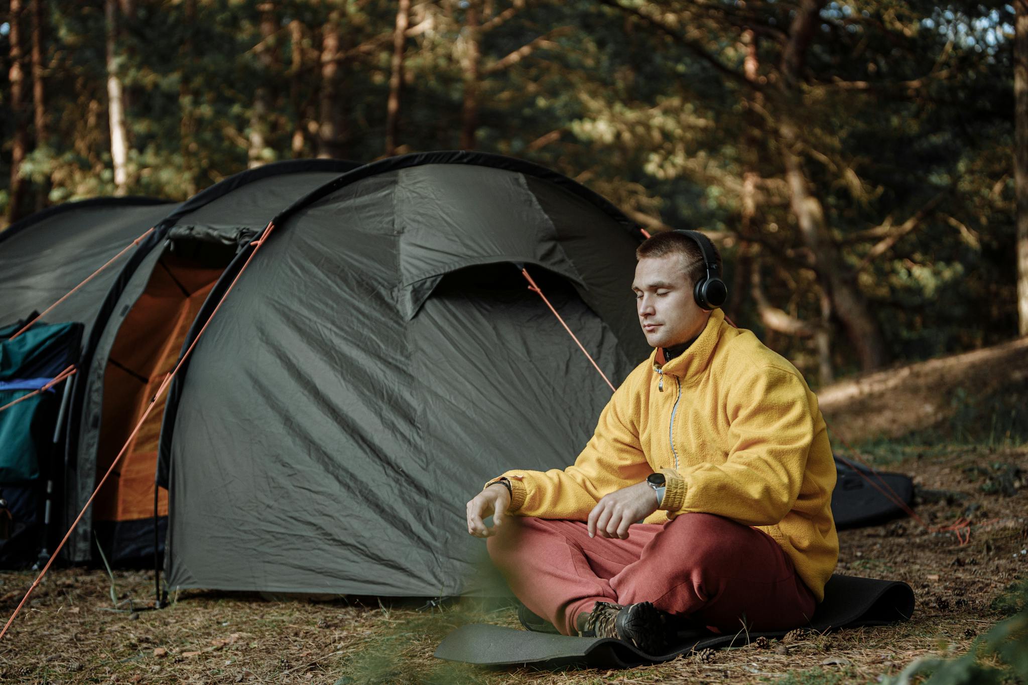 A man enjoys quiet meditation outside a tent in a forest, wearing headphones and a yellow jacket.